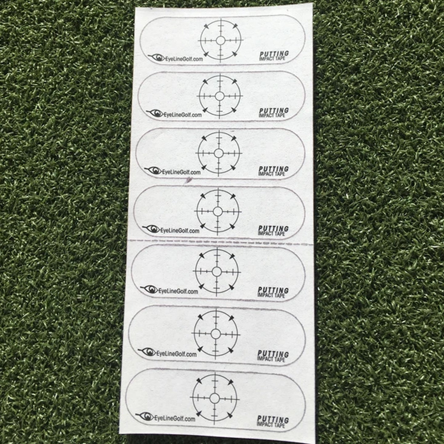 A series of adhesive strips with a target design is laid on artificial grass. Each strip is labeled "PUTTING IMPACT TAPE" and displays a circular crosshair pattern for golf practice.