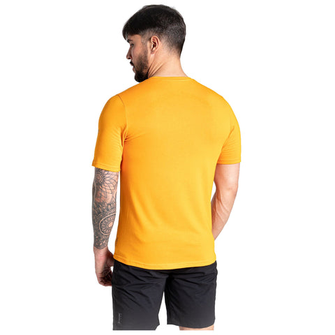 A man with a tattooed arm is wearing a short-sleeved orange shirt and black shorts standing in a neutral background facing slightly away from the camera