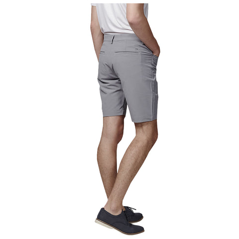 A person stands with their back facing the viewer wearing gray shorts and navy shoes with laces hands in pockets in a neutral setting.