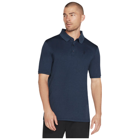 A man stands wearing a navy blue polo shirt with a collar and short sleeves looking slightly to the side in a neutral background suggesting a casual or relaxed setting.