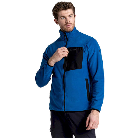 A man is standing while touching the zipper area of a blue fleece jacket with black accents in a neutral background setting, suggesting a casual or outdoor wear context.