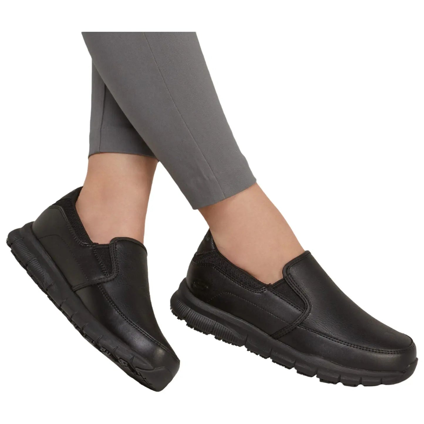 Black slip-on shoes are positioned diagonally, suggesting movement. They are worn with grey fitted pants, set against a plain white background, enhancing the shoe's features.