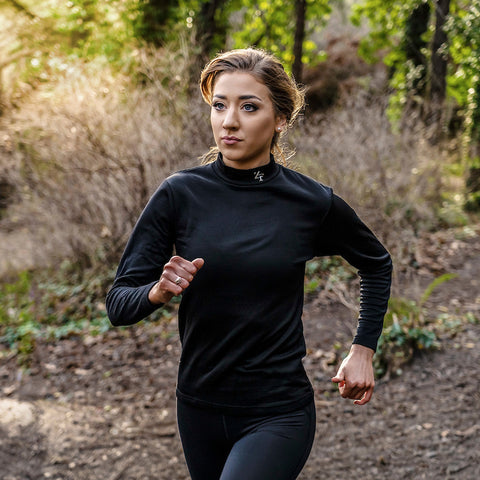 A woman is running along a dirt path through a wooded area wearing a black long-sleeve top and leggings with greenery and bushes in the background.