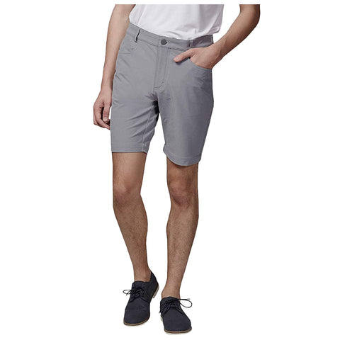 A person stands wearing gray shorts and a white shirt with one hand in a pocket and the other relaxed by their side in a neutral setting.