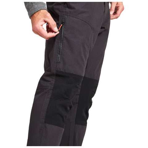 A person adjusts the zippered pocket on the side of dark pants with black paneling while standing in a neutral environment, highlighting the functionality of the clothing.