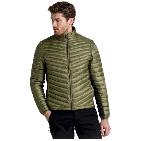 A man wearing a green puffer jacket stands with a serious expression his right hand rested in his pocket while he gazes slightly to his left against a plain background.
