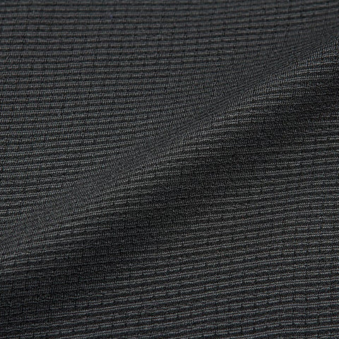 A textured dark fabric lies in folds showcasing a series of fine, parallel ridges creating a subtle pattern against a neutral backdrop suggesting clothing or upholstery fabric in a softer light setting