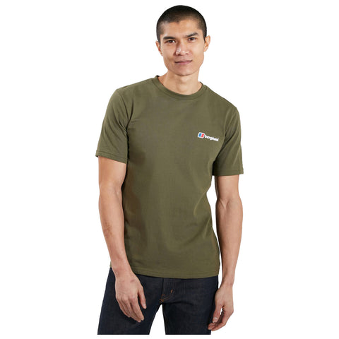 A young man stands confidently wearing an olive green t-shirt featuring a logo on the left side while dark jeans complete his casual outfit against a plain white background.