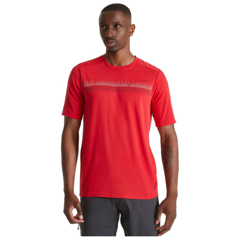 A man stands confidently wearing a bright red short-sleeve shirt featuring a graphic pattern across the chest in a neutral setting with soft lighting.