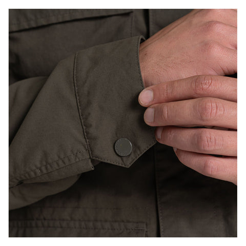 A hand is positioned near the cuff of an olive green jacket while adjusting a snap button on the sleeve. The background is neutral and unobtrusive, focusing on the clothing detail.