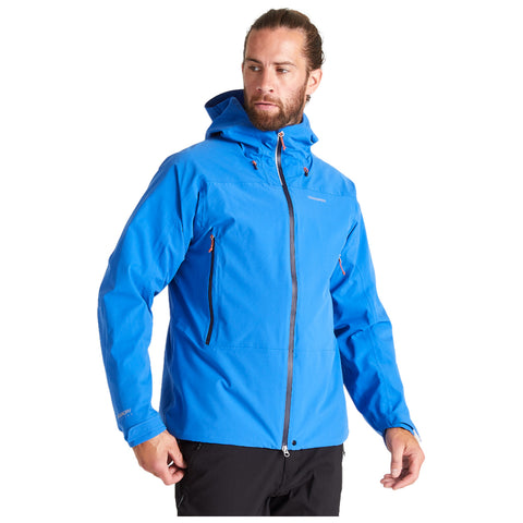A man is wearing a blue jacket with a hood and zippers standing in a neutral setting. He appears to be looking to the side while slightly adjusting the jacket.