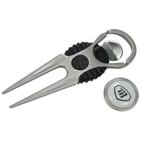 A silver golf tee tool with a forked end rests next to a round ball marker featuring a logo on a white background in a neutral setting.