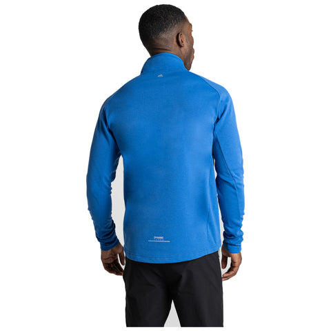 A man is standing with his back to the viewer wearing a blue long-sleeve shirt and black pants in a neutral background setting.