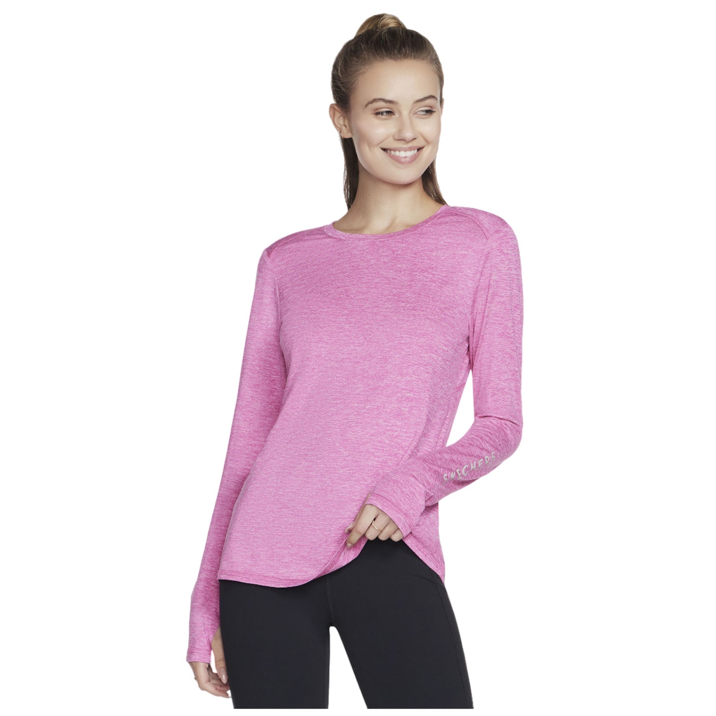A woman smiles while wearing a long-sleeved pink top with a rounded hem and black leggings in a neutral setting, suggesting a casual or athletic context.