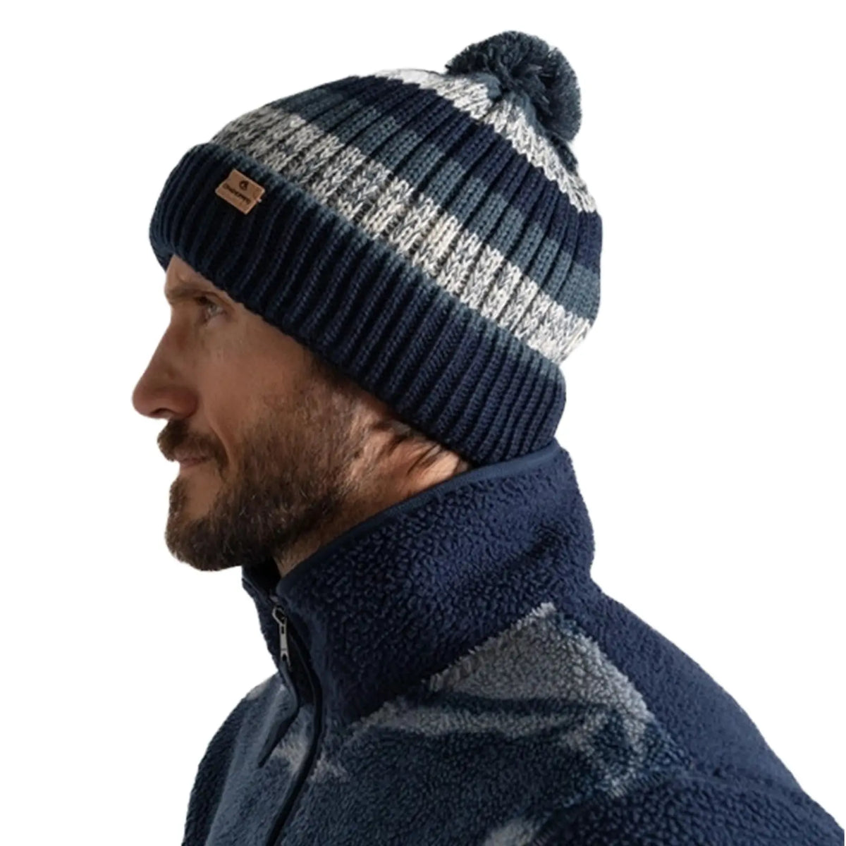 A man is wearing a navy and gray striped knitted beanie with a pom-pom on top standing against a plain light background, dressed in a dark fleece jacket.