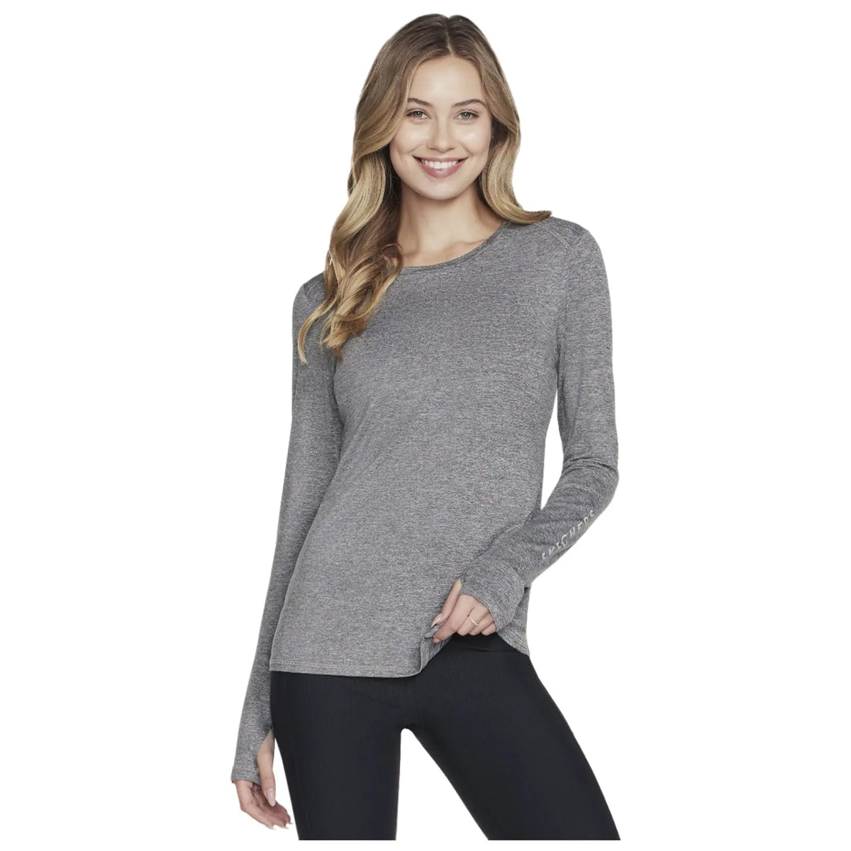 A woman smiles while wearing a fitted long-sleeve gray top paired with black leggings. She poses confidently against a plain background, showcasing a casual yet stylish look.