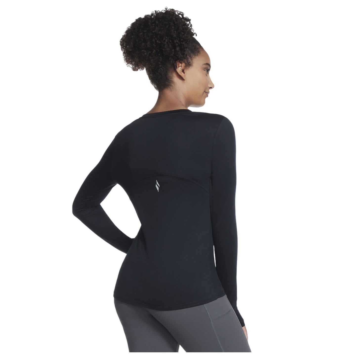 A woman stands with her back to the viewer wearing a fitted long-sleeve black top and gray leggings in a plain white background showcasing her athletic apparel.