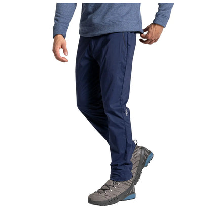 A man stands calmly wearing blue hiking pants and grey outdoor shoes with a textured surface while reaching for the waistband of his pants in a neutral setting.