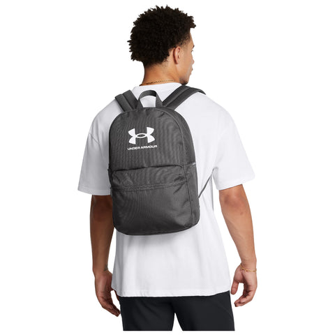 A person wears a black Under Armour backpack while facing away holding the straps in a casual setting wearing a white shirt and dark pants with a simple background.