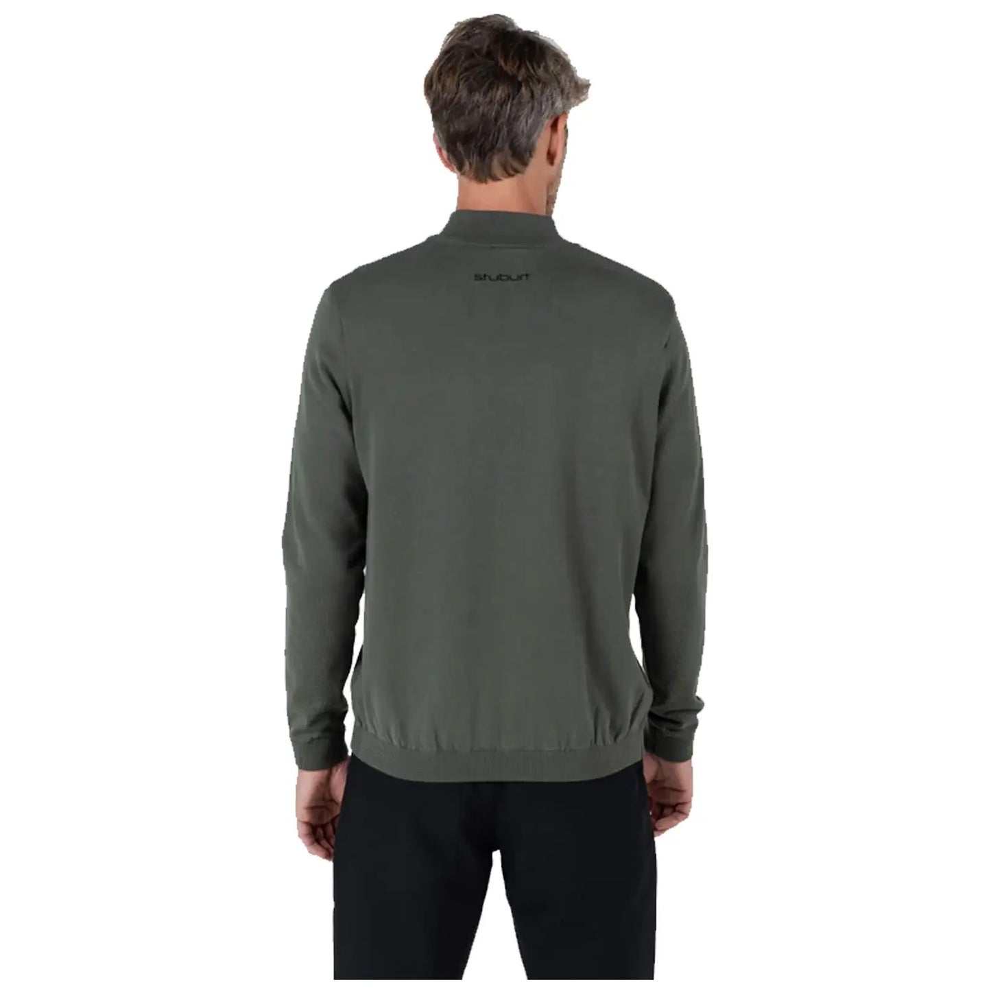A man stands with his back to the viewer wearing a dark green sweater and black pants in a neutral space highlighting the clothing design and fit.