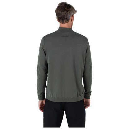 A man stands with his back to the viewer wearing a dark green sweater and black pants in a neutral space highlighting the clothing design and fit.