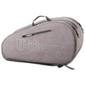 A gray tennis bag with a prominent white Wilson logo is resting on a flat surface featuring multiple compartments and a top handle for carrying.
