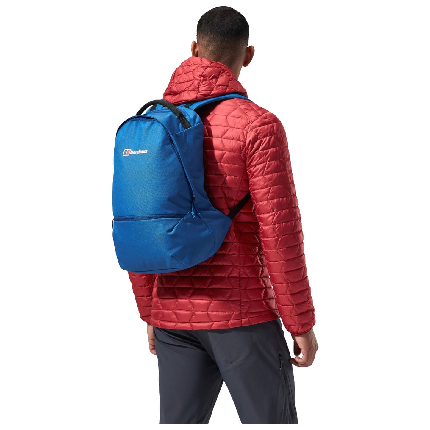 A person wearing a red quilted jacket and dark pants stands with their back to the viewer, carrying a blue backpack with the brand name visible in a plain background.