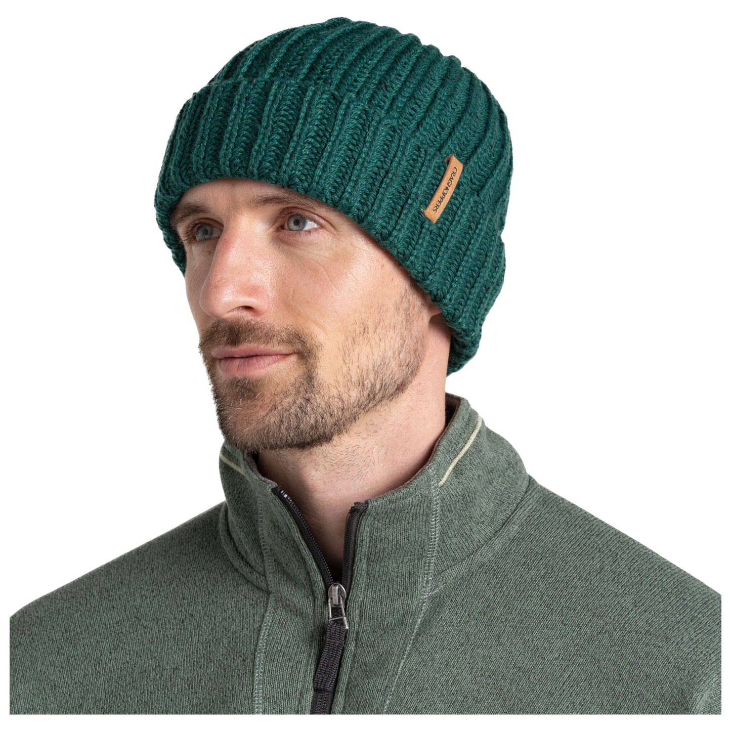 A man wears a green ribbed knit beanie while looking slightly to the side. He is dressed in a gray zippered jacket and appears calm in a neutral background.