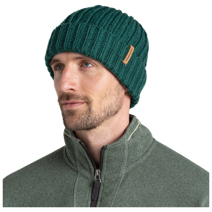 A man wears a green ribbed knit beanie while looking slightly to the side. He is dressed in a gray zippered jacket and appears calm in a neutral background.