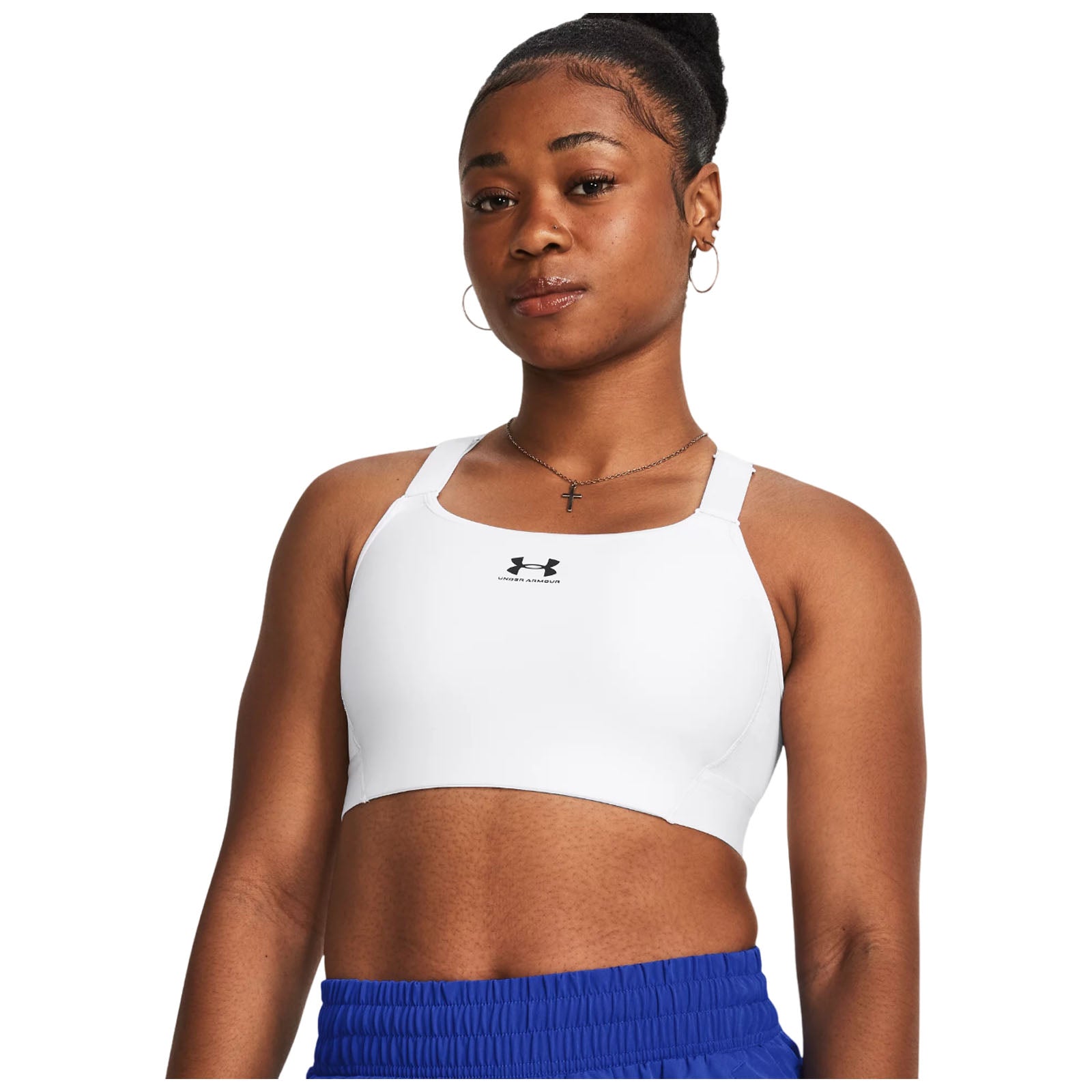A young woman stands confidently wearing a white athletic crop top with a logo and blue shorts in a bright, neutral setting, showcasing a determined expression and stylish accessories.