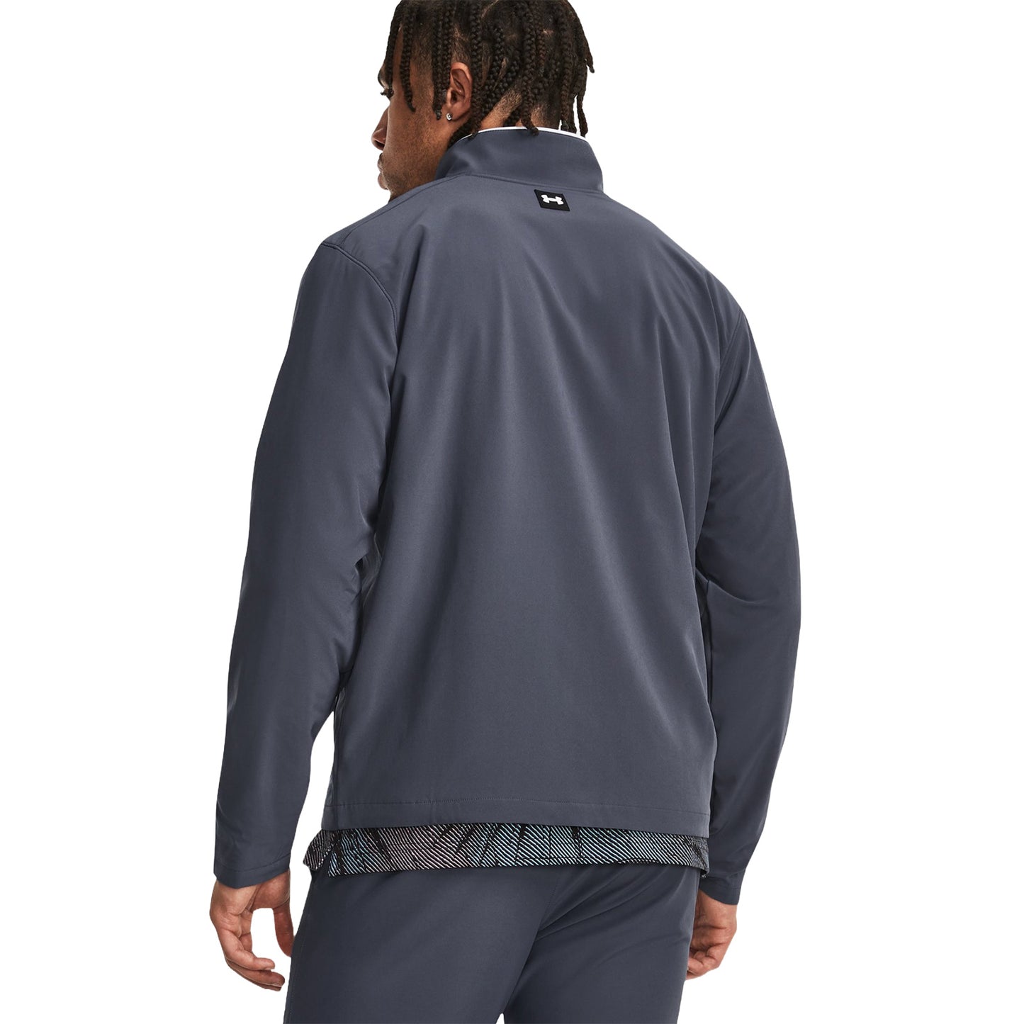 A man wearing a dark gray athletic jacket stands with his back to the viewer showing part of a patterned shirt beneath in a plain white background.