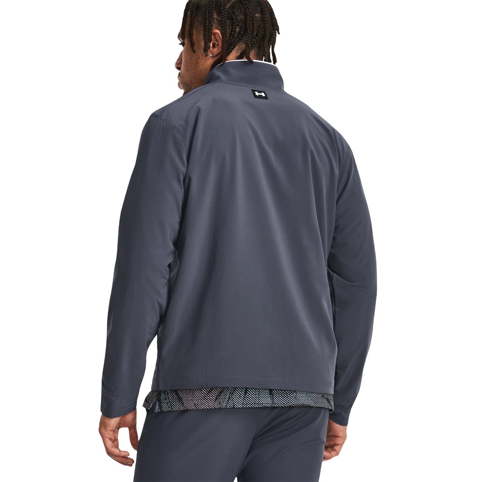 A man wearing a dark gray athletic jacket stands with his back to the viewer showing part of a patterned shirt beneath in a plain white background.