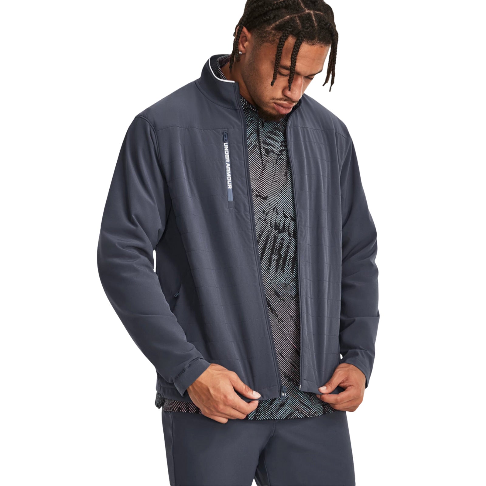 A man wearing a dark gray athletic jacket and patterned shirt is looking down at his hands while standing against a plain white background. His braided hair is visible.