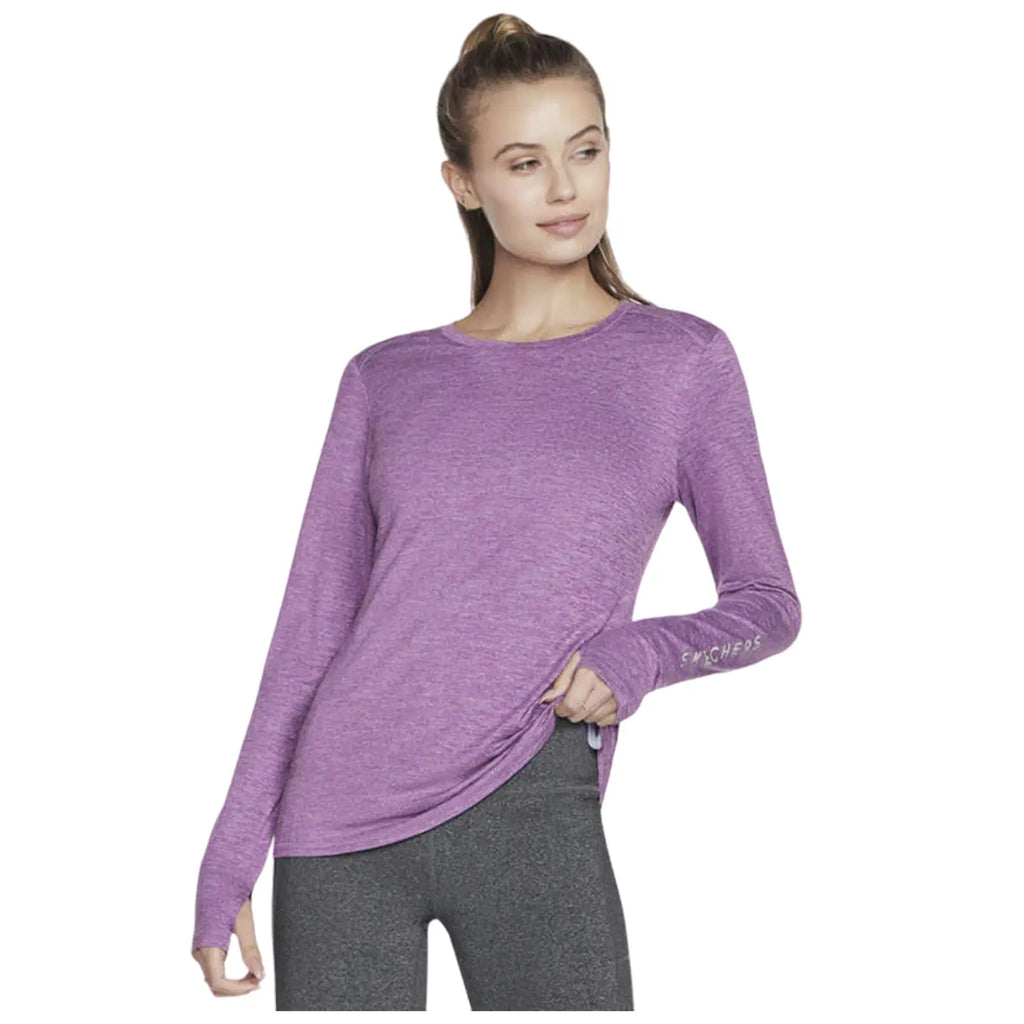 A woman wears a long-sleeve purple top and gray leggings. She stands confidently with a slight smile and a hand on her hip in a neutral background setting.