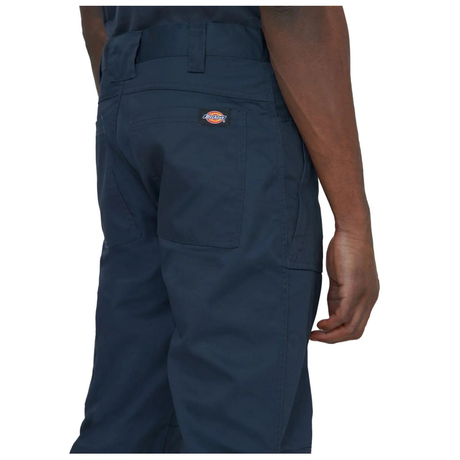 A person stands facing away highlighting dark blue pants with a logo patch their hand rests at their side in a neutral setting against a plain background