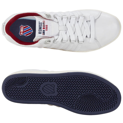 A pair of white athletic shoes is displayed with one shoe inverted to show the navy blue sole while revealing the brand logo and year on the inside of the other shoe.
