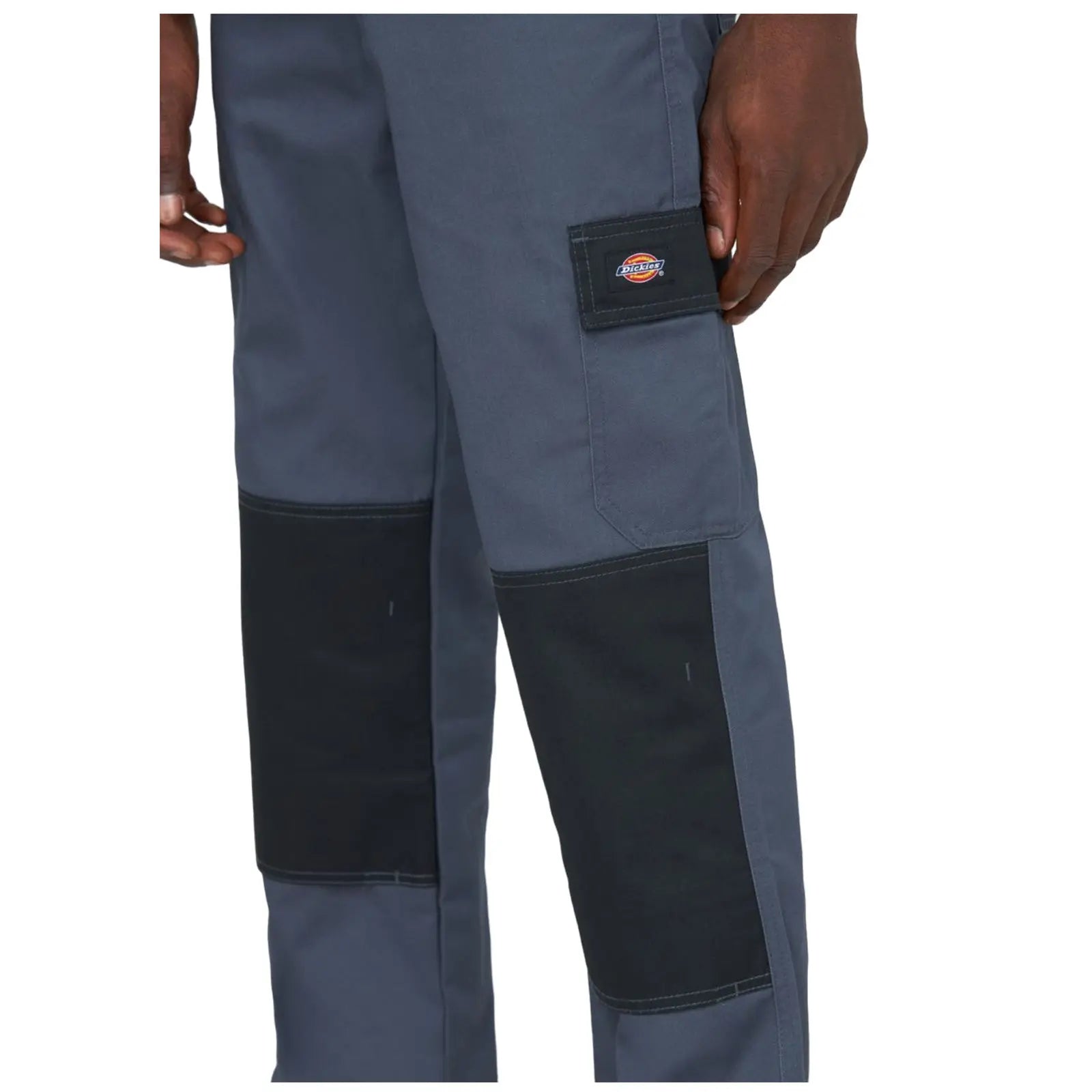 Gray work pants with black reinforced patches on the knees are being held by a hand. The pants feature a small logo on the side pocket.