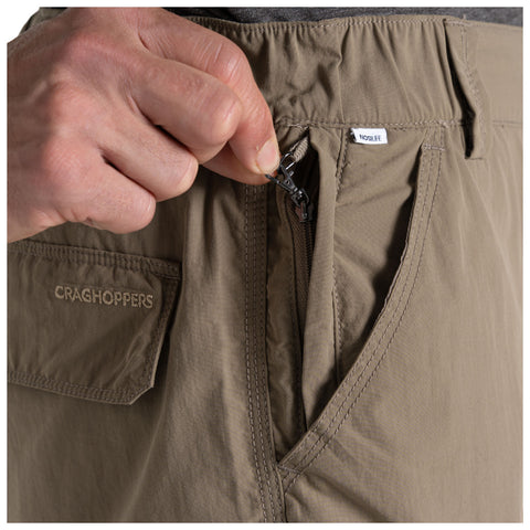 A hand is gripping a zipper while pulling it up on a pair of brown Craghoppers shorts, showcasing a close-up view of the clothing item and its fastening mechanism.