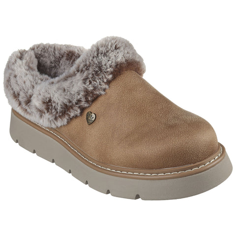 A tan slipper with a soft, fluffy interior lining rests on a flat surface featuring a small heart-shaped logo on the side providing warmth and comfort for indoor use