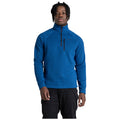 A person stands confidently wearing a blue fleece pullover with a half-zip collar and black pants in a neutral background. They have shoulder-length curly hair.