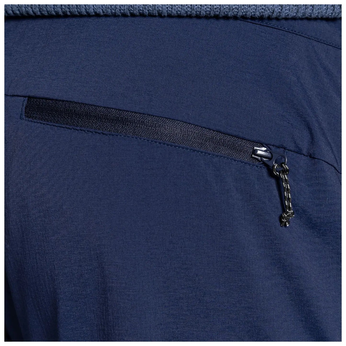 A close-up of a dark blue fabric with a zippered pocket on the right side featuring a small pull cord on the zipper revealing the texture of the material around it.