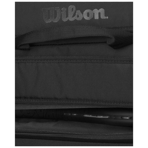 A black bag with a visible Wilson logo lies open displaying its interior contents including a tennis racket featuring a black handle and strings in a dark environment.