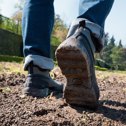 A muddy shoe steps forward in soft earth with jeans rolled up nearby in a sunny outdoor setting surrounded by greenery and trees in the background
