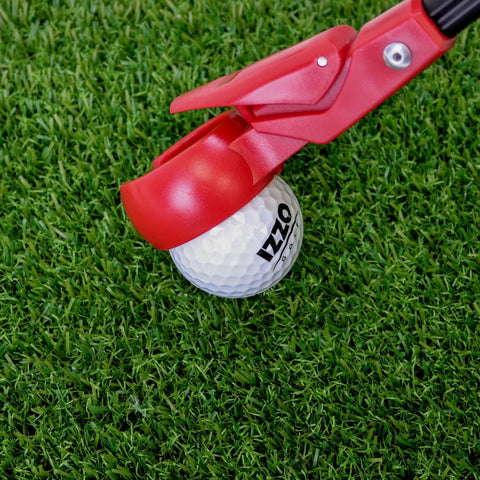 A red golf ball holder is positioned above a white golf ball marked with IZZO on artificial green grass, preparing to assist in teeing the ball for a shot.