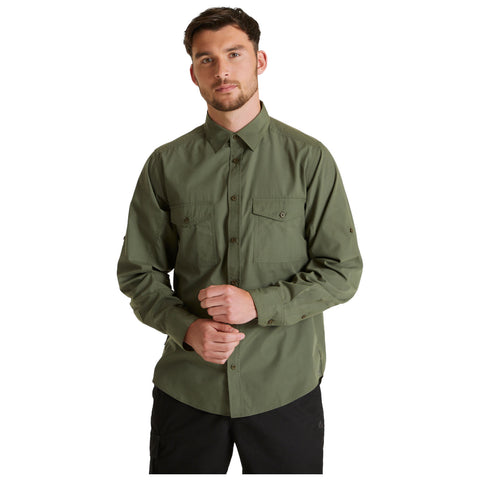 A man wears a green long-sleeved shirt with buttoned pockets and stands with his hands together in front of him against a plain white background.