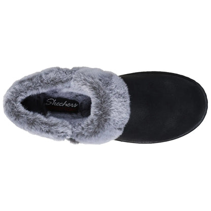 A black slipper with a gray fur lining rests on a flat surface showcasing its cozy design and branding that reads Skechers Memory Foam inside the opening.