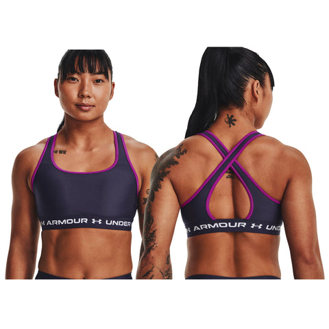 A young woman stands confidently, wearing a bright pink sports bra with a black band displaying "UNDER ARMOUR." She has long, dark, wavy hair and a neutral expression.