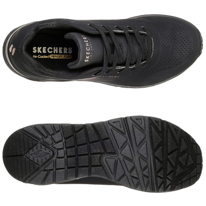 Black sneakers are positioned side by side with one foot slightly raised showcasing their design while a person wearing olive green pants stands in a neutral setting highlighting the footwear.