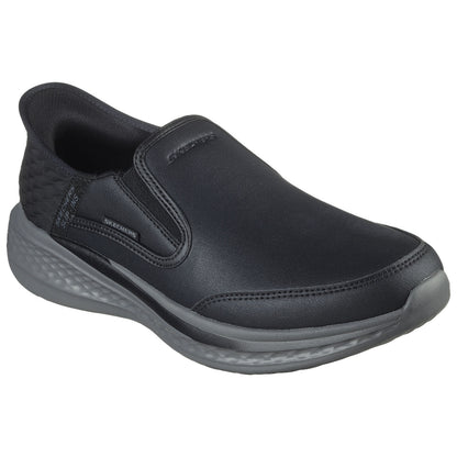A black slip-on shoe features a smooth leather upper and elastic side panels for easy wear resting on a textured gray sole in a minimalistic setting.