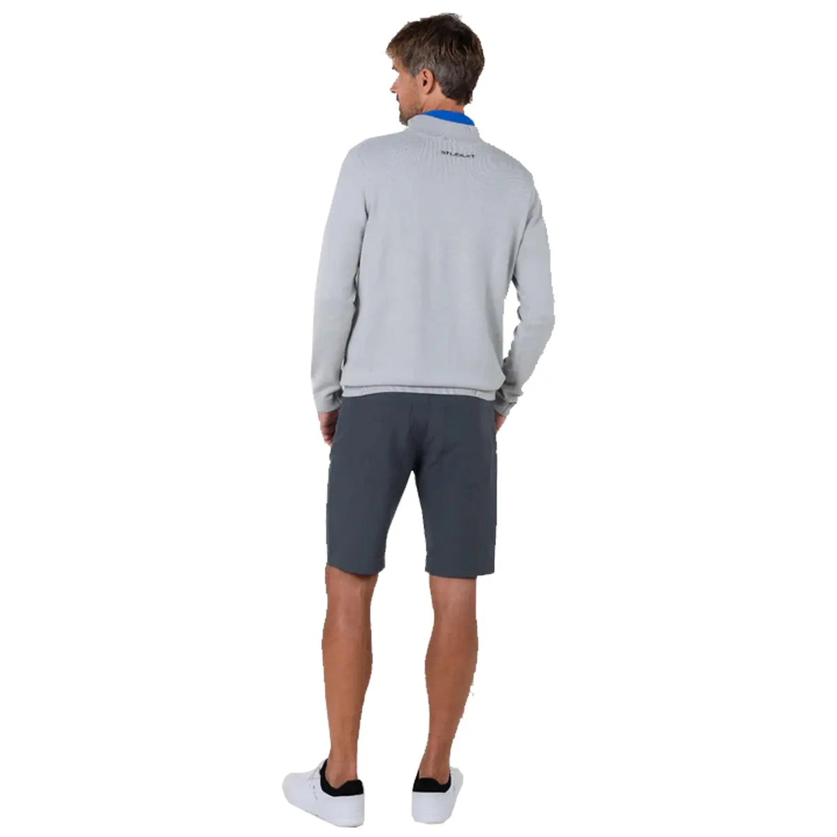 A man stands with his back turned wearing a grey sweater and dark shorts in a neutral backdrop, suggesting a casual, relaxed environment appropriate for outdoor activities.
